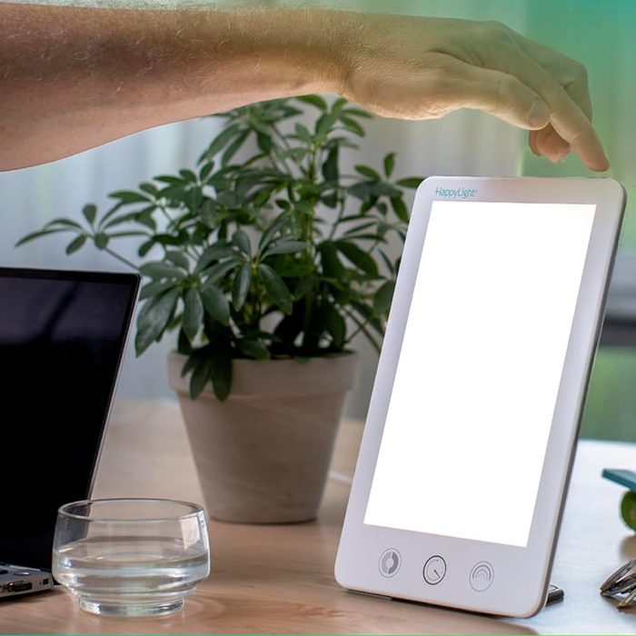 Light therapy lamp, touched by a hand, sits on a desk near a laptop, glass of water, and potted plant.