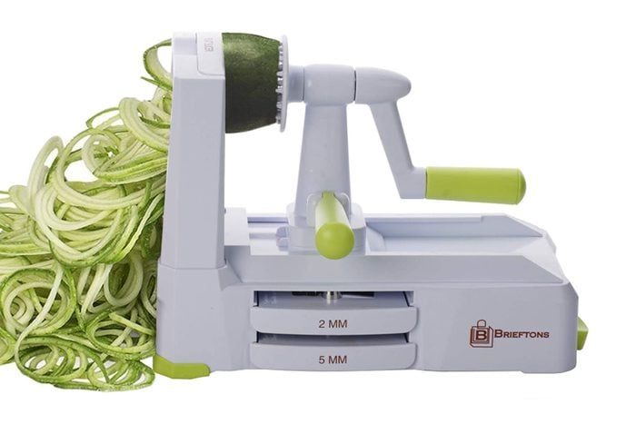 A vegetable spiralizer creates zucchini noodles on a white surface; the machine features green accents and labeled blade settings of 2mm and 5mm.