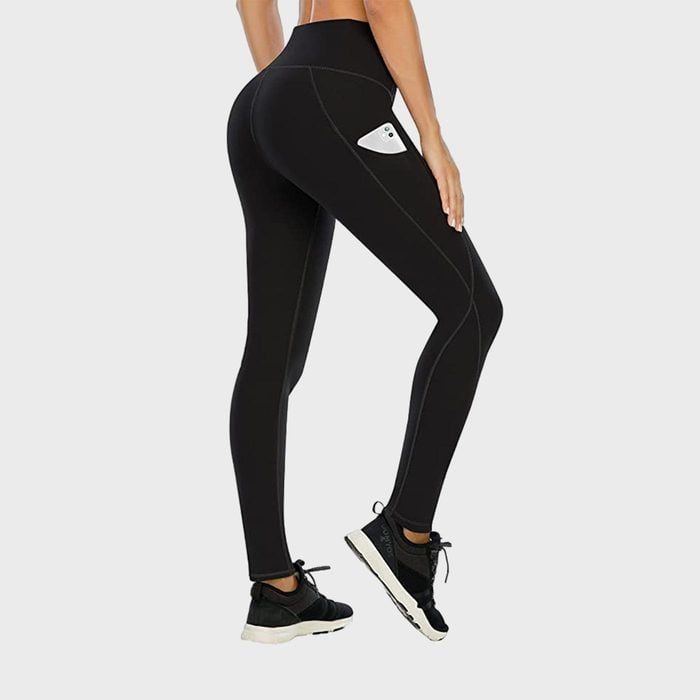 A person wears black leggings with a phone in the pocket, standing indoors against a plain background, wearing black sneakers.