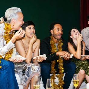 People sitting, eating popcorn, and wearing gold decorations; appear joyful in a festive indoor setting with champagne glasses nearby.