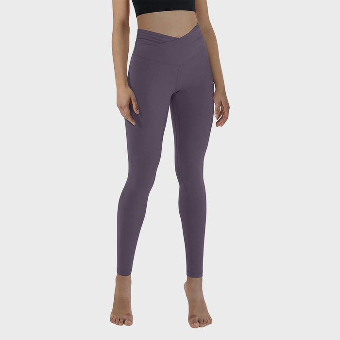 Person wearing high-waisted, purple leggings and a black crop top, stands barefoot on a plain white background.