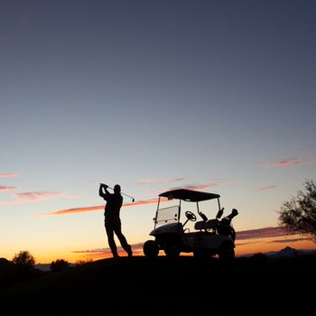 Silhouette of golfer swinging a club next to a golf cart at twilight with a mountain and tree in the background