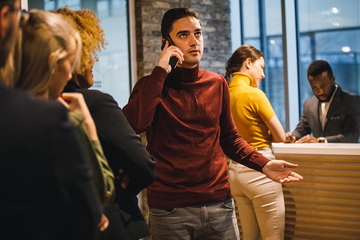 Man in line to check in at a hotel holds a phone up to his ear with an exasperated expression